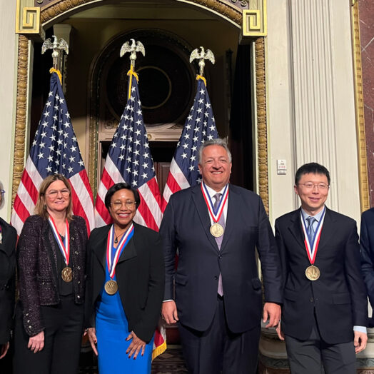 Arati Prabhakar, Angela Belcher, Paula Hammond, Noubar Afeyan, Feng Zhang, and Emory Brown pose together in front of 3 U.S. flags.